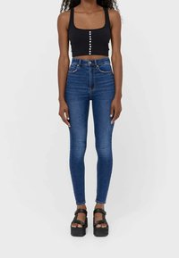 Svart kort topp med fyrkantig halsringning och knapppdetalj; högmidjade blå denim skinny jeans; svarta platåsandaler.