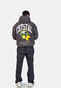 Felpa grigia con cappuccio, con la scritta "CRYSTAL" stampata in bianco sul retro, caratterizzata da una grafica di limone e fiori. Abbinata a jeans scuri e sneakers bianche.