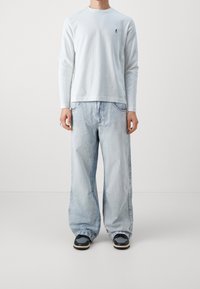 Vaaleansininen pitkähihaisten paita, jossa on pieni logo rinnassa, yhdistettynä väljät vaaleansinisiin farkkuihin ja tummiin lenkkareihin. Yksinkertainen, rento muotoilu.