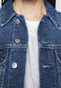 Jeansjacka i blått, med silverlogo-knappar, strukturerad krage och framfickor. Bärs över en vit t-shirt. Texturerad tyg.