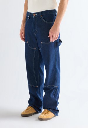 Hombre vistiendo jeans de mezclilla azul oscuro y holgados con múltiples bolsillos y zapatillas color beige, de pie frente a un fondo blanco liso.