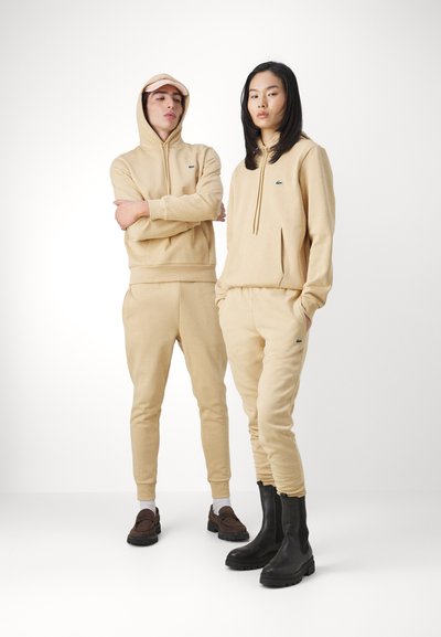 Ensemble de sweat à capuche beige et de pantalon de jogging, avec une poche kangourou et un petit logo. Le modèle porte des mocassins marron et des bottines noires.texture uniforme.