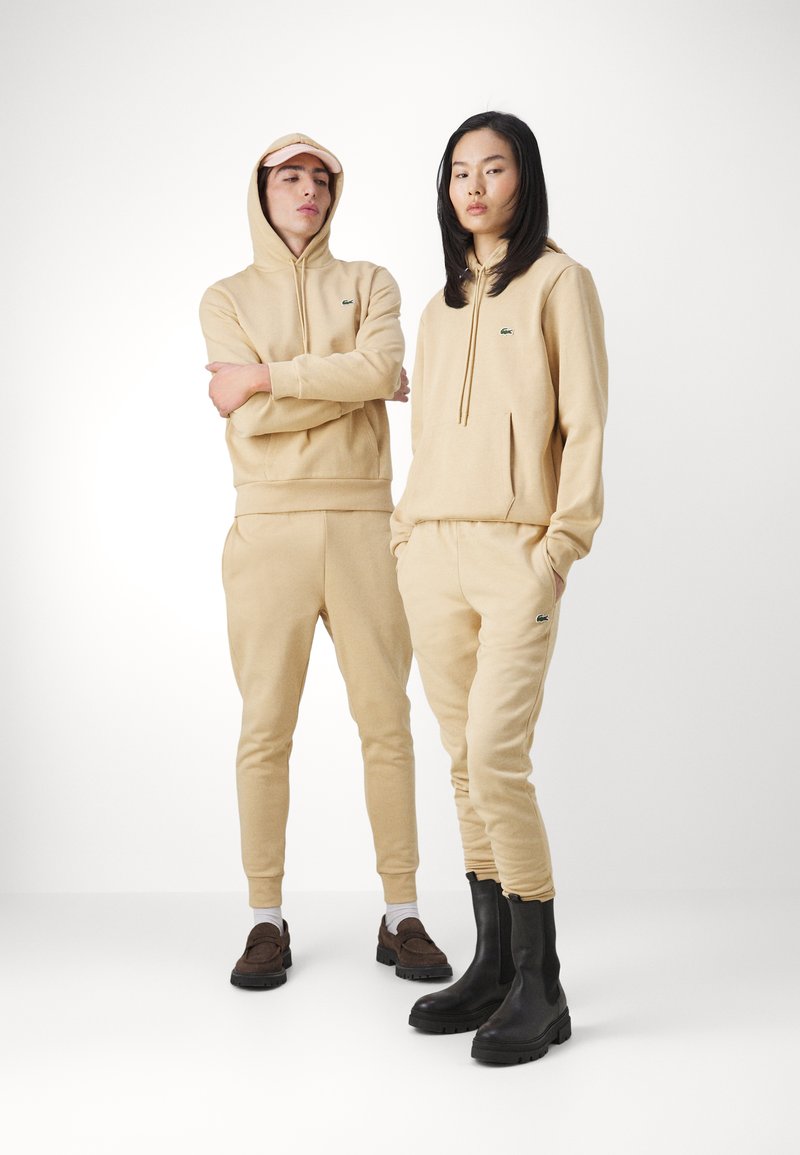 Beige Kapuzenpullover und Jogginghosen-Set mit Kängurutasche und kleinem Logo. Das Modell trägt braune Slipper und schwarze Stiefeletten. Einheitliche Textur.