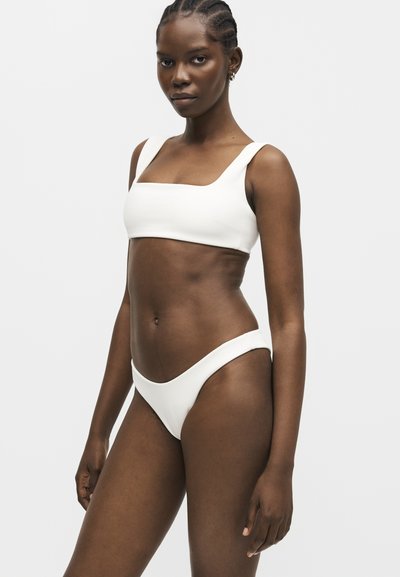 Femme aux cheveux tressés portant un haut de bikini blanc à encolure carrée et un bas de bikini taille haute assorti sur un fond clair uni.