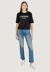 Maglietta nera a maniche corte con logo "GUESS" bianco, abbinata a jeans azzurri e sneakers bianche. Design casual e semplice.