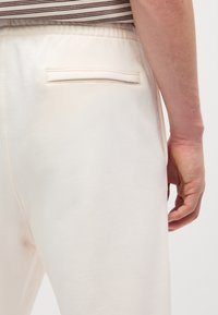 Kremowe dresy z tylną kieszenią, o gładkiej strukturze i elastycznym pasku. Częściowo widoczna jest w paski koszula.