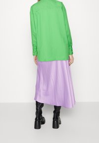 Grüne Hemdbluse, lilafarbenen Maxirock, schwarze Stiefeletten mit Absatz. Glatte Texturen, kontrastierende Farben und entspannter Schnitt mit sichtbaren Nähten und Akzenten.