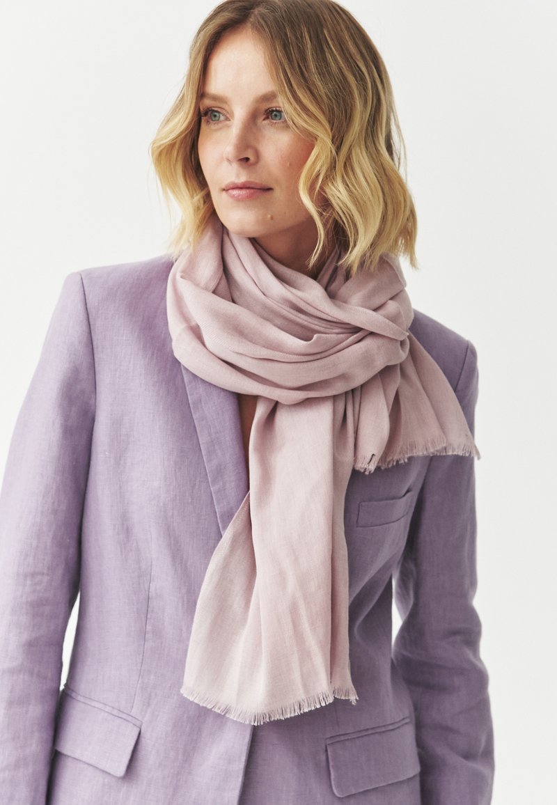 Ljusrosa halsduk i ett mjukt, strukturerat tyg draperar runt halsen, tillsammans med en lila kavaj med notch-krage och ficka.