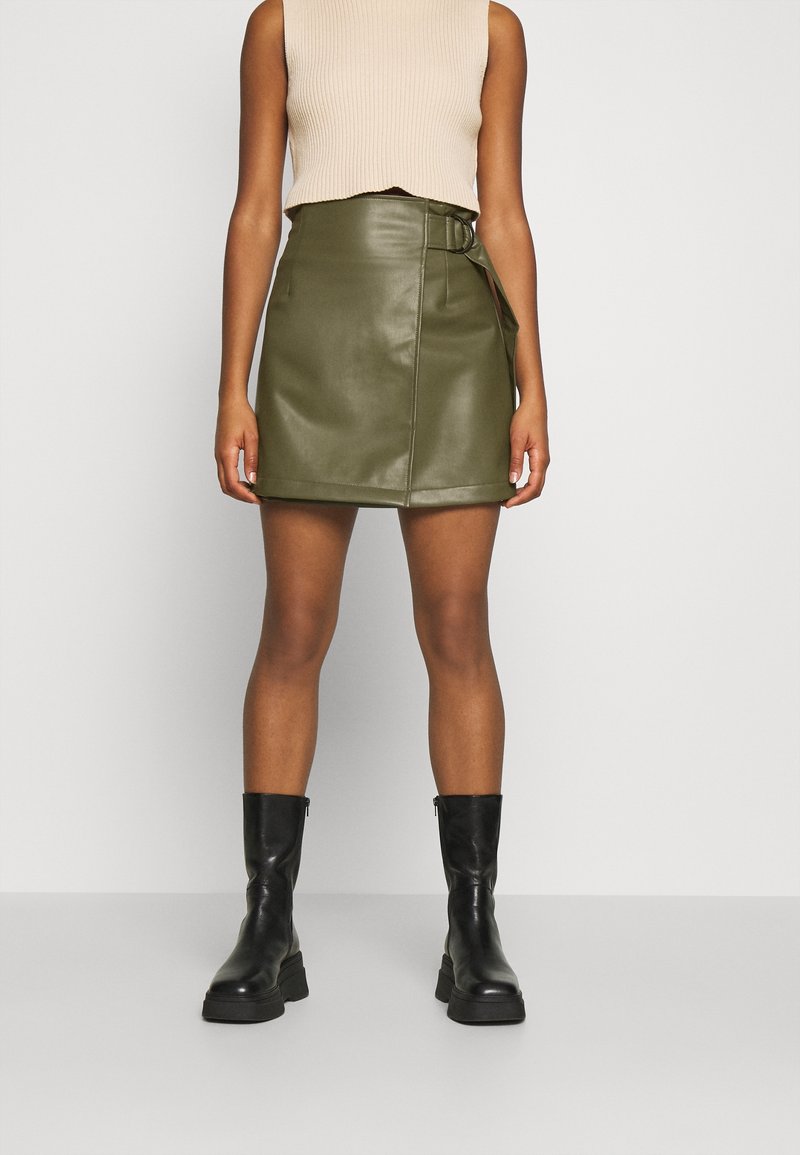 Jupe mini en simili cuir olive avec un design asymétrique et un détail de lien sur le côté. Portée avec un haut en maille beige et des bottines noires.
