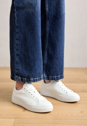 Personne portant un jean bleu à jambes larges et des baskets blanches basses, debout sur un sol en bois.