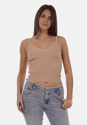 Giovane donna con lunghi capelli castani che indossa un top a maglia beige senza maniche e jeans blu chiaro, in piedi con una mano in tasca.