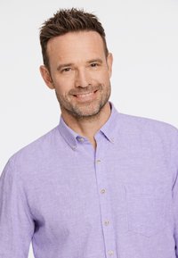 Camisa de botones de color púrpura claro hecha de una tela texturizada, con un bolsillo en el pecho y pequeños botones redondos. Presenta un cuello con solapa.