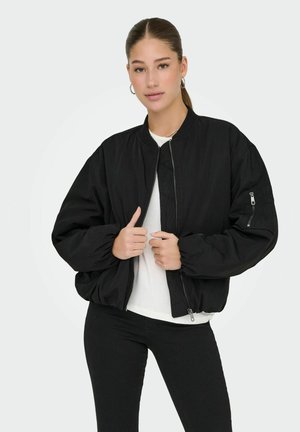 Crna bomber jakna s patentnim zatvaračem sprijeda, oversized kroj, rebraste manžete i džep s patentnim zatvaračem na lijevom rukavu. Nosena preko bijele majice.