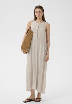 Frau trägt ein ärmelloses beiges Leinenkleid und braune Flip-Flops, hält eine große hellbraune Wildledertasche vor weißem Hintergrund.