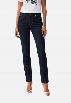 Donna che indossa jeans blu scuro a gamba dritta, tacchi bianchi aperti sulle dita e una t-shirt bianca con stampa colorata di pappagallo.