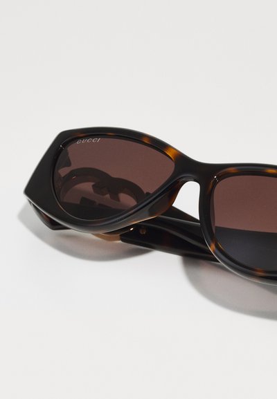 Occhiali da sole tartarugati con un design geometrico, lenti marroni scure e il logo "Gucci" stampato sulla lente superiore sinistra.