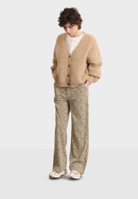 Cardigan beige lavorato a maglia con scollatura a V, bottoni in legno; abbinato a pantaloni color sabbia con stampa leopardata nera, indossato con sneakers chiare.