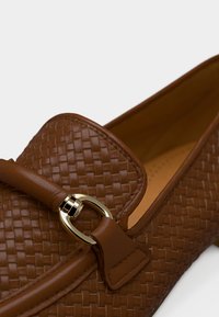 Καφέ υφαντά δερμάτινα μοκασίνια με στρογγυλεμένη μύτη, που διαθέτουν διακοσμητικό αγκράφα σε χρυσό τόνο και λείο δέρμα στην επάνω επιφάνεια.