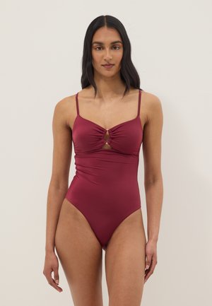 Donna con lunghi capelli scuri indossa un costume intero bordeaux senza maniche con dettagli a ritaglio sulla parte anteriore, in piedi davanti a uno sfondo chiaro e semplice.