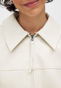 Kremo spalvos dirbtinės odos striukė su dideliu apykakle, priekiniais užtrauktukais ir siuvinėjimu; glotni tekstūra ir minimalistinis dizainas.