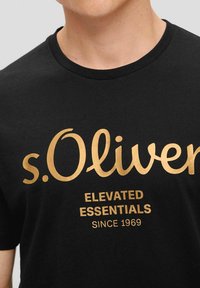 Black cotton t-shirt featuring gold text that reads "s.Oliver" with "ELEVATED ESSENTIALS" and "SINCE 1969" below in a bold font.