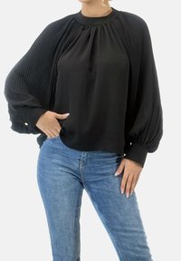 Zwarte blouse met geplooide mouwen, ronde halslijn en knoopmanchetten. Draagt bij blauwe skinny jeans. Gladde stof, losse pasvorm.
