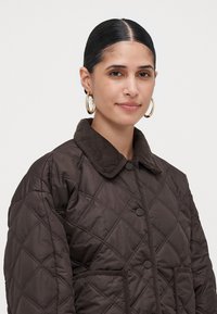 Woman with slicked-back hair wearing gold hoop earrings and a dark brown quilted jacket with corduroy collar and snap buttons.