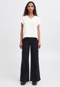Top bianco a V con maniche corte abbinato a pantaloni neri a vita alta e a gamba larga. Scarpe nere completano l'outfit. Sfondo semplice.