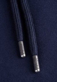 Close-up of two navy blue braided hoodie drawstrings with metal tips engraved "SPITZBUB" on dark blue fabric.