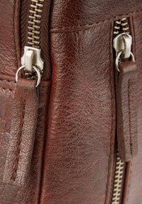 Détail d'un sac en cuir marron montrant une fermeture à double zip avec quincaillerie argentée, surface texturée et languettes en cuir.