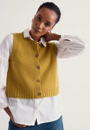 Femme portant un gilet en maille jaune moutarde sur un chemisier blanc et un jean bleu, une main posée sur son cou, tournée vers l'avant.