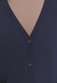 Mörkblå cardigan med V-ringning, ribbad textur och en enda brun knappstängning framtill. Tyget verkar mjukt och stretchigt.