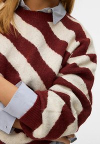 Rot-weiß gestreifter Pullover mit strukturiertem Strick, ausgestattet mit einem gerippten Rundhalsausschnitt und sichtbaren, geschichteten blauen Hemdmanschetten darunter.