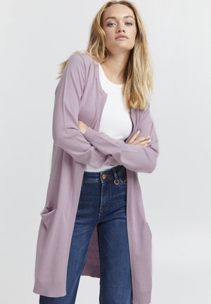 Cardigan lilas à ouverture devant, avec des poignets côtelés et deux poches à l'avant, associé à un haut blanc et un jean bleu. Tissu lisse et léger.