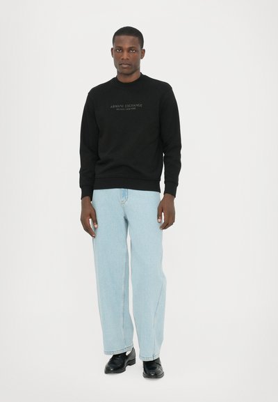 Sweatshirt noir avec des poignets côtelés, doté d'un logo "Armani Exchange" en relief. Associé à un jean bleu clair et des chaussures noires.