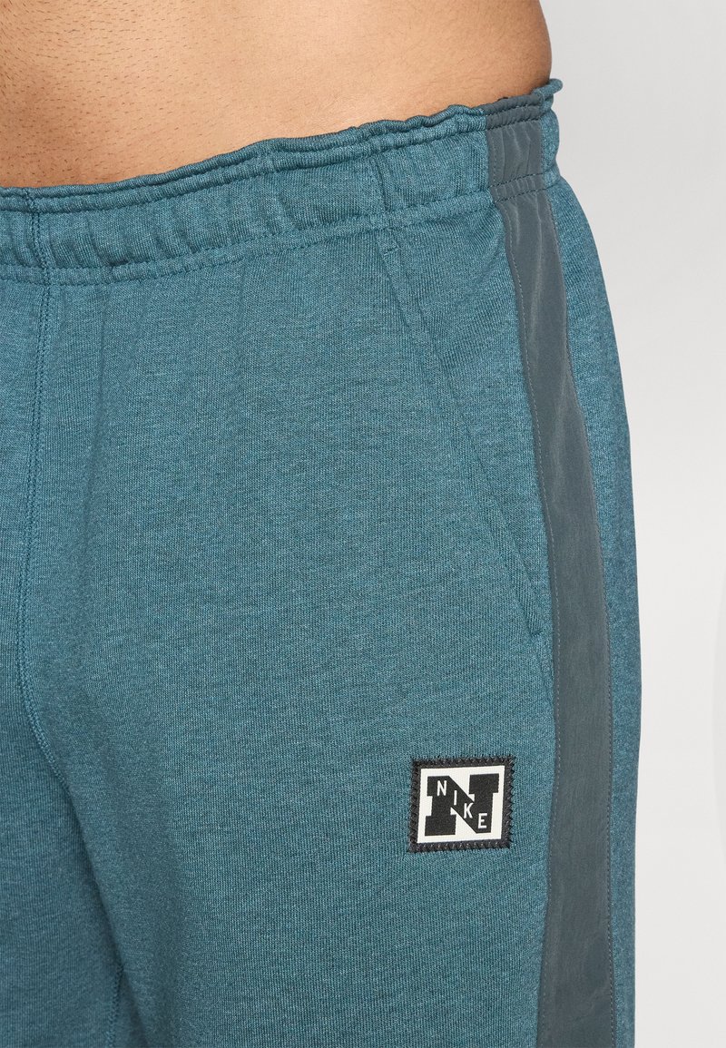 Teal joggebukser lavet af blødt stof, med elastisk talje, sidelommer i en mørkere nuance og et sort Nike-logo. 