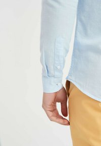 Chemise à manches longues bleu clair avec des poignets à bouton, offrant une coupe décontractée et une texture lisse. Associée à un pantalon beige, la main reposant sur le côté.