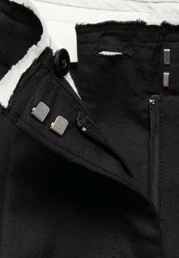 Tissu noir avec un gros plan sur une fermeture éclair, deux attaches crochets métalliques, un bouton et une boutonnière sur les bords du vêtement.