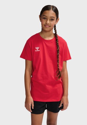 T-shirt en coton rouge avec un col rond et des manches courtes, arborant un petit logo blanc sur la poitrine gauche. Porté avec un short noir.