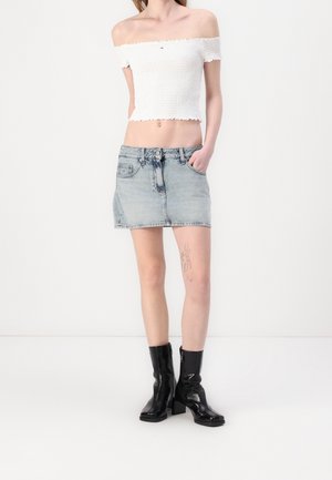 Person trägt ein weißes schulterfreies Crop-Top, einen hellblauen Jeans-Minirock, schwarze Stiefeletten mit Absatz und eine sichtbare Oberschenkel-Tätowierung.
