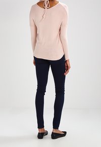 Ljusrosa tröja med rundad halsringning och knytning bak, kombinerad med mörkblå slim-fit byxor och svarta ballerina-skor.