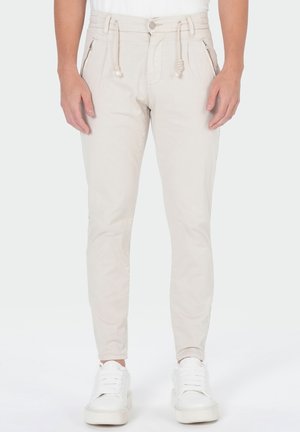 Beige slim-fit broek met ritszakken en koordriem, gedragen met witte sneakers en een wit topje, getoond van taille tot voeten.