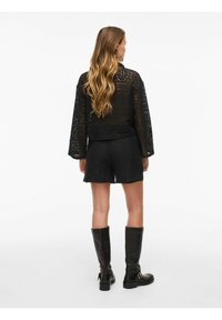 Czarna koronkowa bluzka z długimi rękawami, zestawiona z czarnymi szortami w wzory. Całość dopełniają czarne kozaki za kolano. Pokazano widok z tyłu.