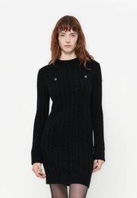 Abito nero lavorato a maglia con texture a coste, girocollo e maniche lunghe. Presenta bottoni decorativi sulle spalle e polsini a coste.