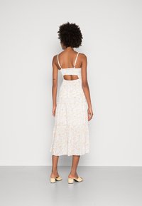 Weißes Blumenmaxikleid mit verstellbaren Trägern, gerafftem Rücken und Cut-Out-Detail. Weicher Stoff fällt bis zu einem stufigen Saum. Kombiniert mit beigen Blockabsatz-Mules.