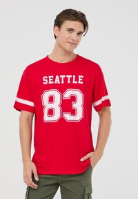 T-shirt in cotone rosso con maniche corte, caratterizzata da testo bianco "SEATTLE" e grafica "83". La maglietta ha un fit rilassato e strisce a contrasto.