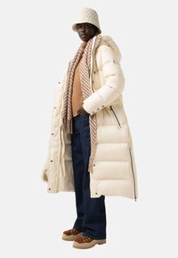 Lang, beige pufferjakke med hætte, parret med en tan sweater og stribet tørklæde. Mørkeblå brede jeans og tan snørede støvler afslutter looket.