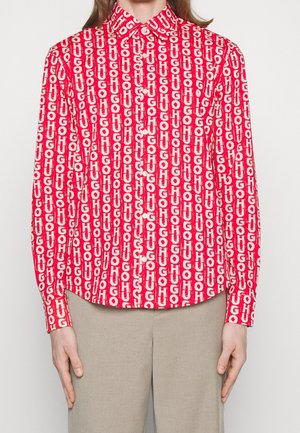 Osoba nosząca jaskrawoczerwoną koszulę na guziki z białym pionowym wzorem tekstu "HUGO", w połączeniu z beżowymi spodniami, ręce swobodnie opuszczone wzdłuż ciała.