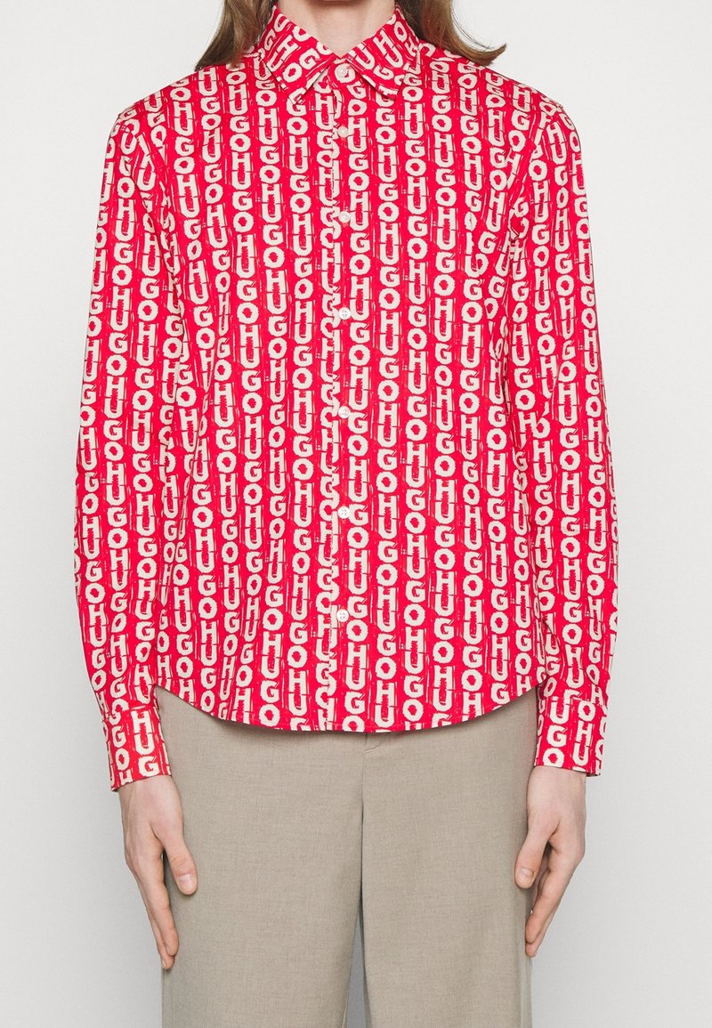 Personne portant une chemise boutonnée rouge vif avec un motif de texte vertical blanc "HUGO", associée à un pantalon beige, mains détendues le long du corps.