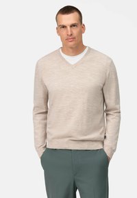 Maglione beige a V in tessuto lavorato, abbinato a una t-shirt bianca sotto, indossato con pantaloni verdi scuri.